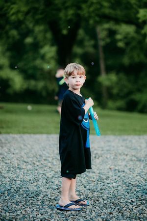 A young child in a black dress and blue flip flops stands on a gravel path with greenery in the background.