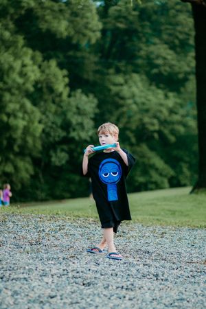 Child blowing bubbles while wearing black shirt with blue graphic design stands on gravel path near wooded area.