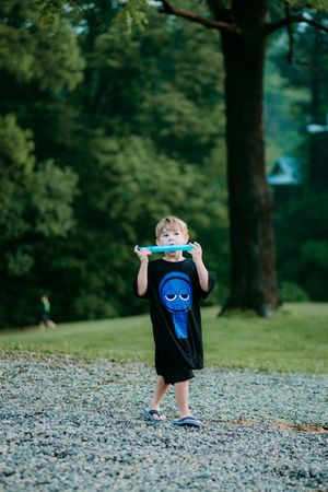 A young child in a black t-shirt with a blue design stands on gravel while playing outdoors.