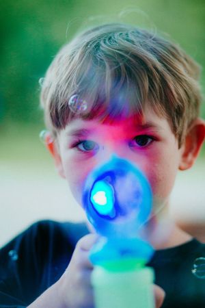 A child blowing into a colorful light-up toy emitting vibrant pink, red and blue illumination against a green background.
