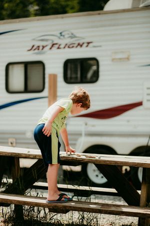 A young child in shorts climbs on a picnic table near a Jay Flight camper trailer at a campground.