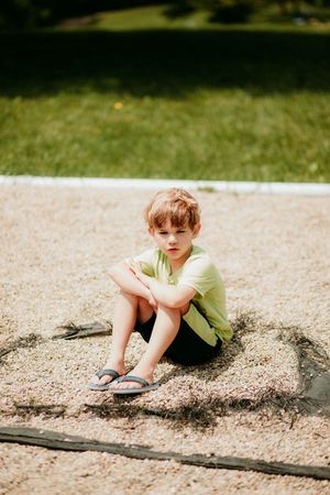 Small child in a yellow t-shirt sits thoughtfully alone on sandy ground on a sunny summer day.