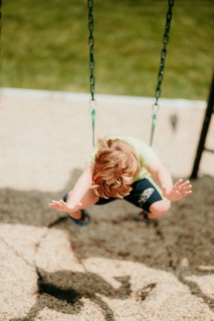 Child in navy blue outfit swings high at a playground, captured in motion against blurred ground shadow.
