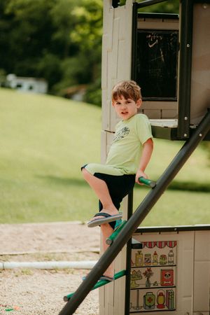Young child in yellow shirt and black shorts climbing on playground equipment in a sunny outdoor setting.