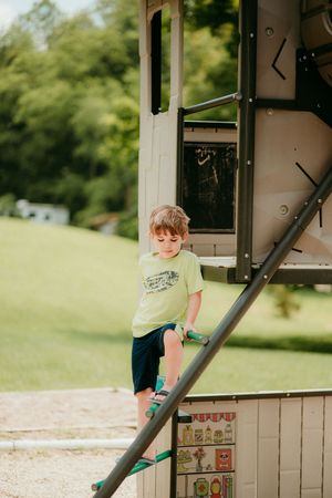 Young child in a green t-shirt climbing on outdoor playground equipment on a sunny summer day.