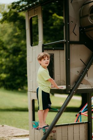 A child in a yellow shirt and shorts plays on a playground structure on a sunny day in a grassy outdoor setting.