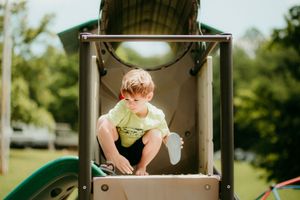 Small child in a lime green shirt sits perched at the top of a playground climbing structure on a sunny day.