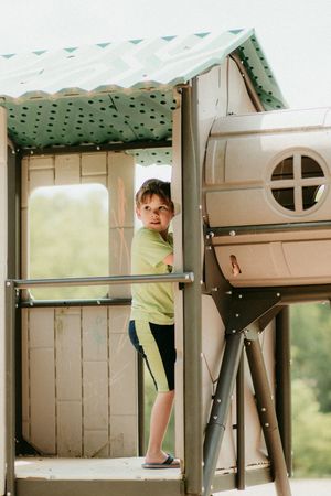 A wooden playground structure with green roofing tile where a child plays inside the elevated tower compartment.