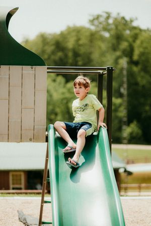 A small child sits at the top of a green metal playground slide on a summer day.