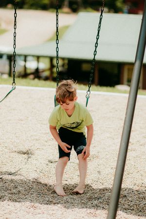 A child in a yellow t-shirt bends down near empty swings in a playground with gravel underneath.