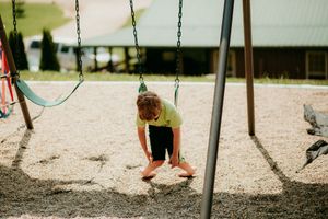 A young figure in a yellow top and black shorts plays on a swing set in a sunny playground area.