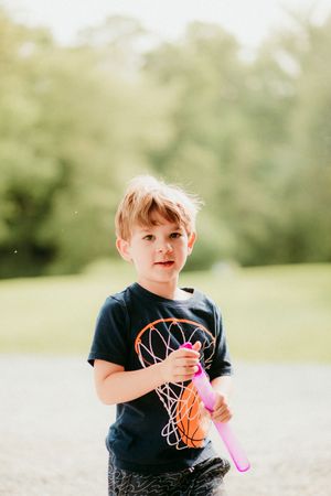 Child in a navy basketball t-shirt holds a pink bubble wand while playing outdoors on a sunny day.