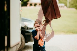 Young person joyfully jumping while holding a red blanket outdoors on a sunny day.