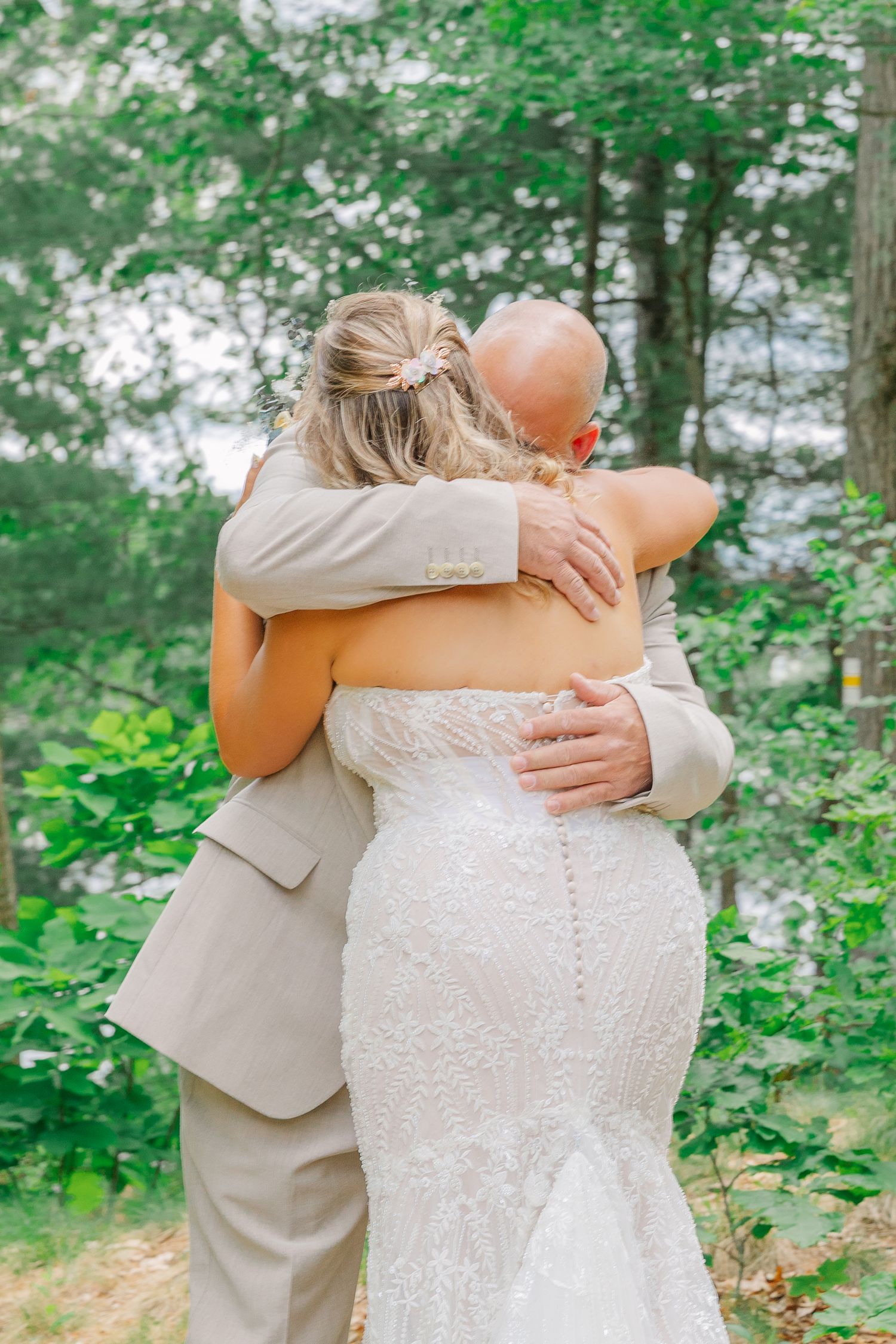 Two people embrace in a romantic wedding moment, wearing a white lace gown and beige suit amid lush green forest surroundings.