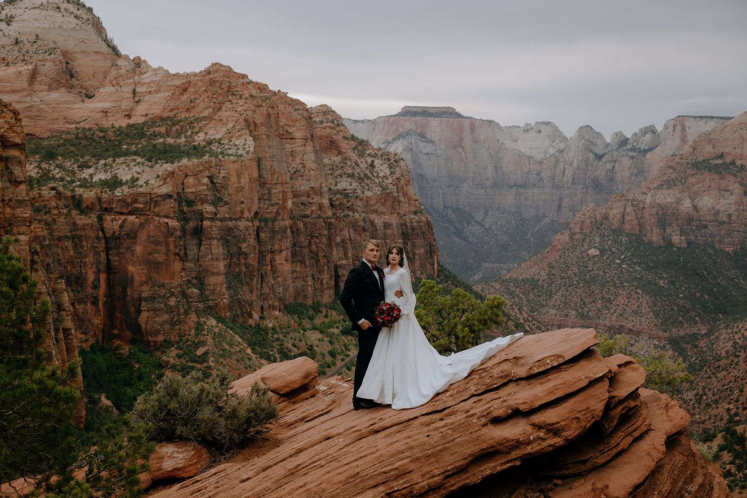 Couple in formal attire stands on red rock overlook with dramatic canyon vista of Zion National Park in background.