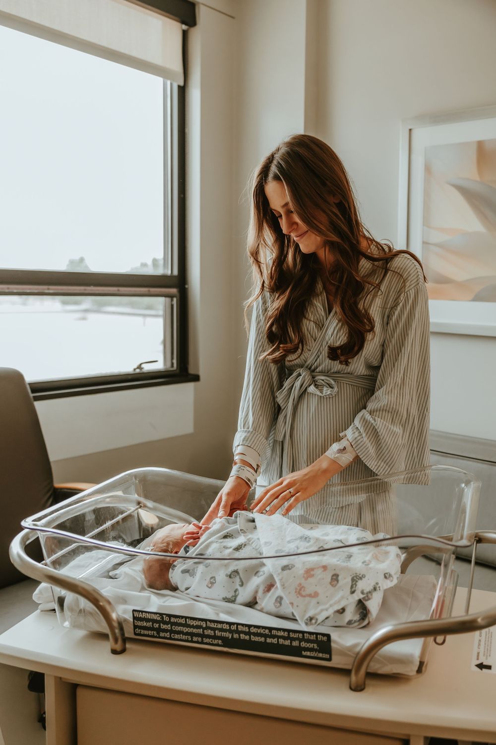 Someone folds baby clothes and blankets on a table next to a bright window.