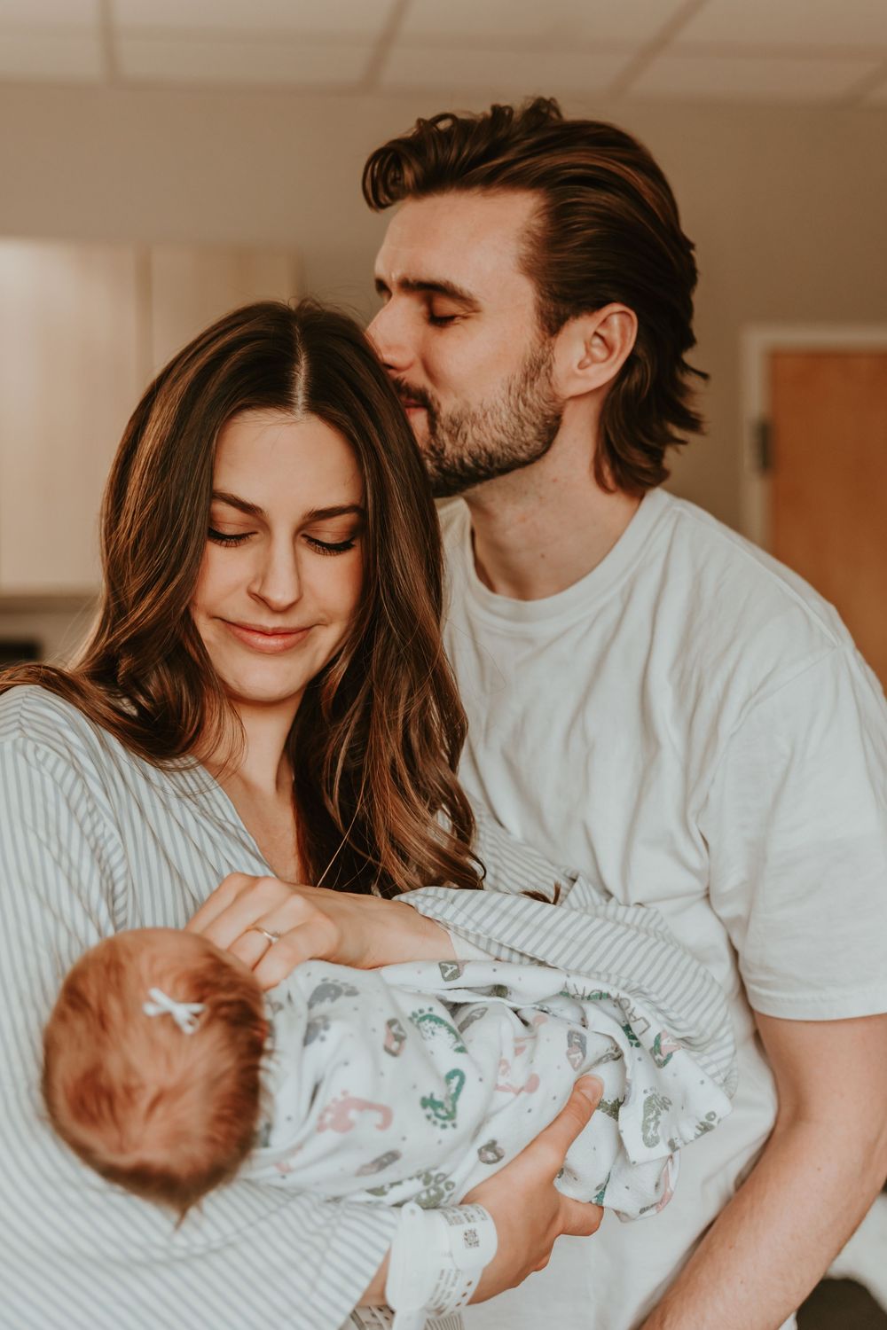 Tender family moment as new parents lovingly cradle and gaze at their sleeping newborn wrapped in a patterned swaddle.