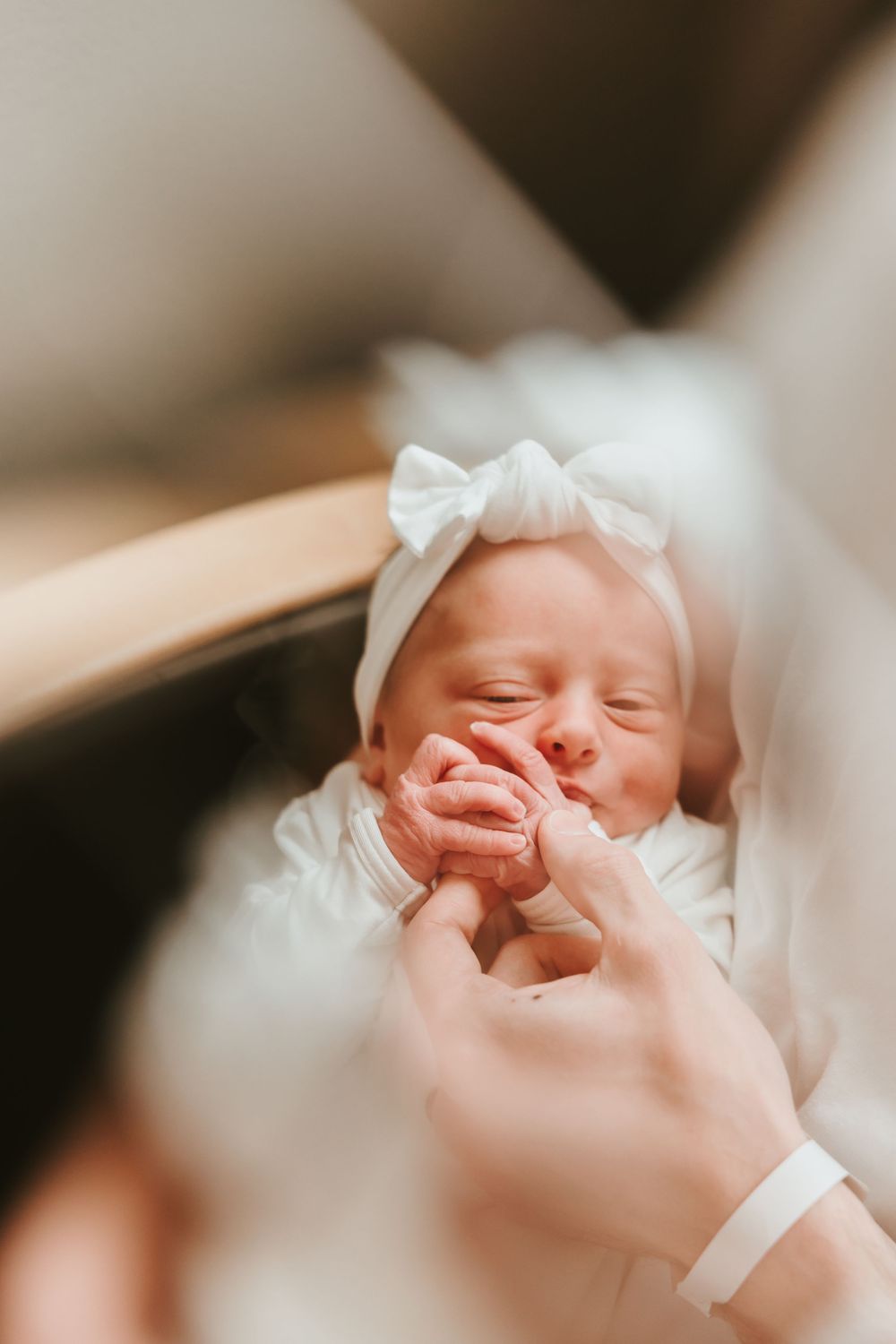 Sleeping newborn wearing white bow headband being gently held by caring hands.