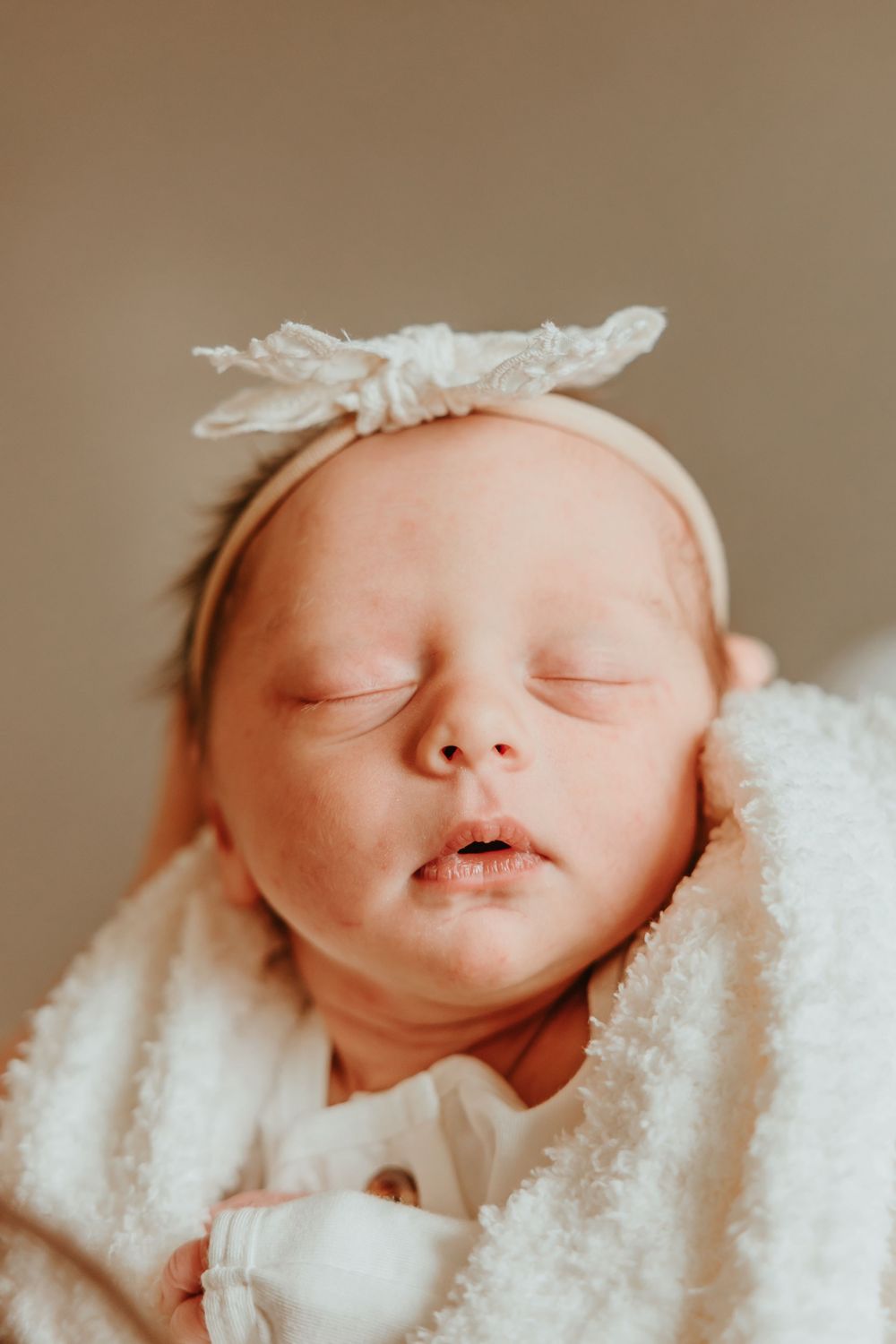 Peaceful portrait of sleeping newborn wearing lace headband and wrapped in soft white blanket.