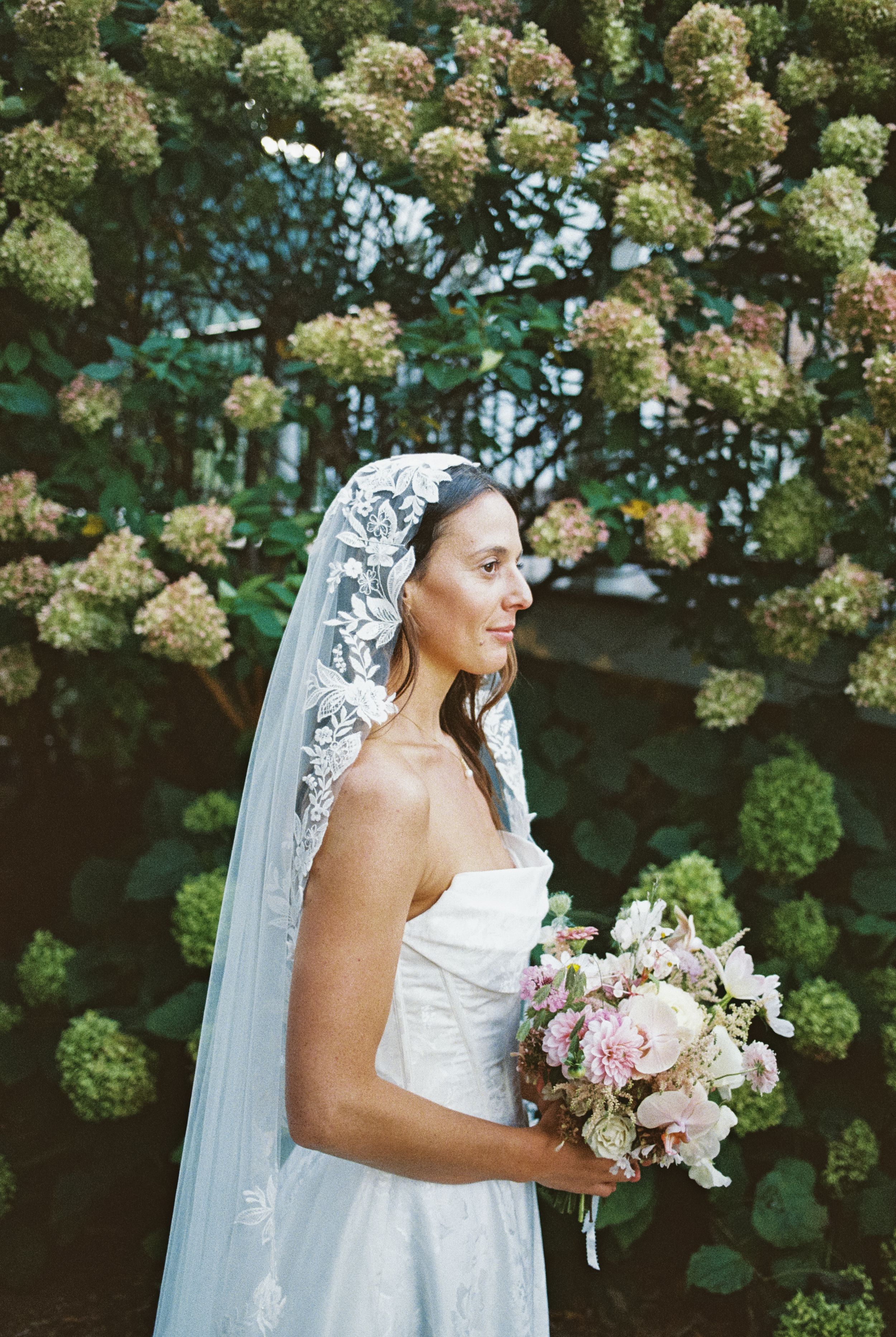 A bride in white dress holding a pink and blush bouquet stands in front of lush hydrangea bushes during a garden wedding.