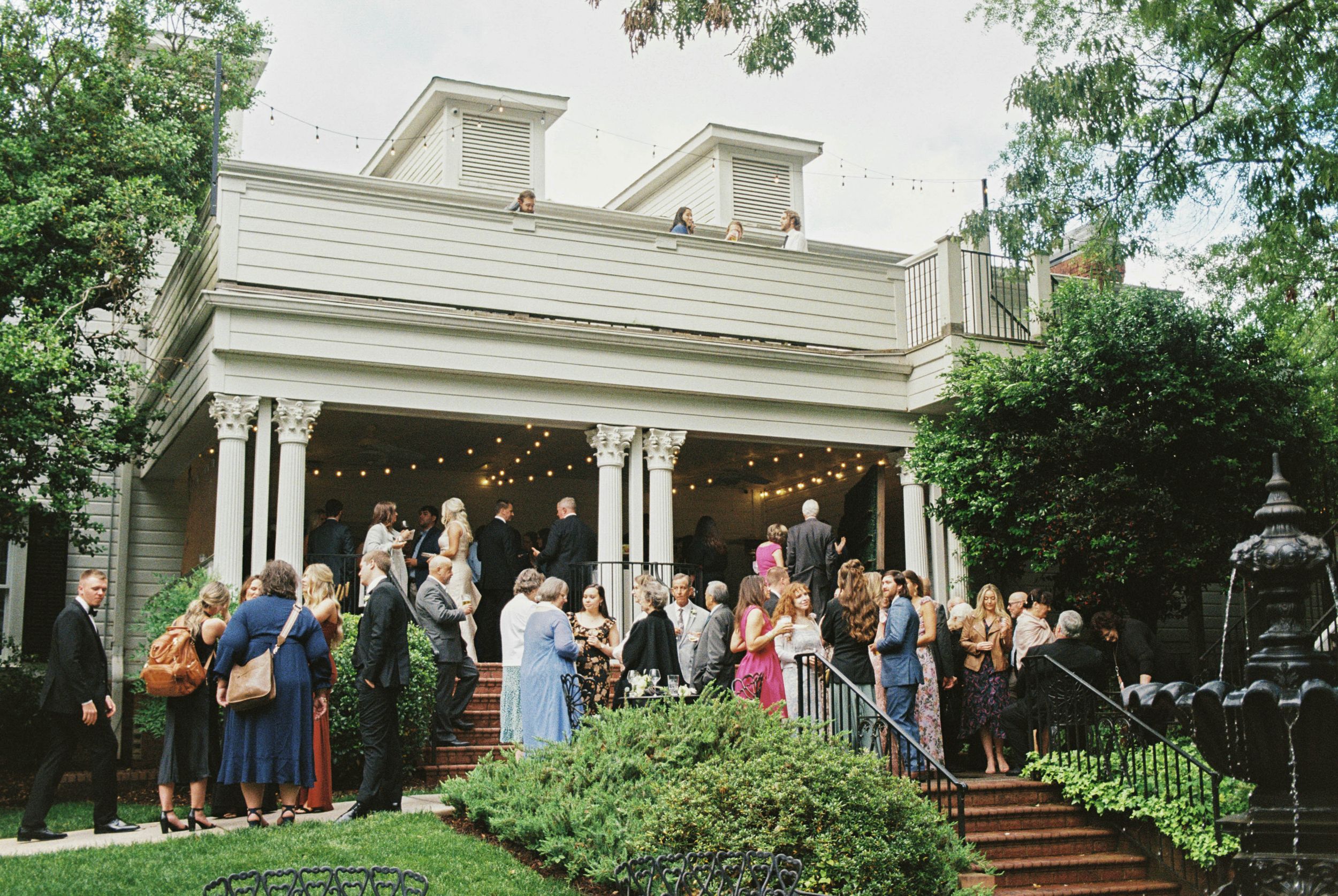 A group of people gather on the steps of a historic white house with dormers and a covered porch in springtime.