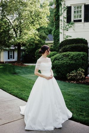 A bride in an elegant white ball gown stands on a concrete path beside a manicured lawn and traditional house exterior.