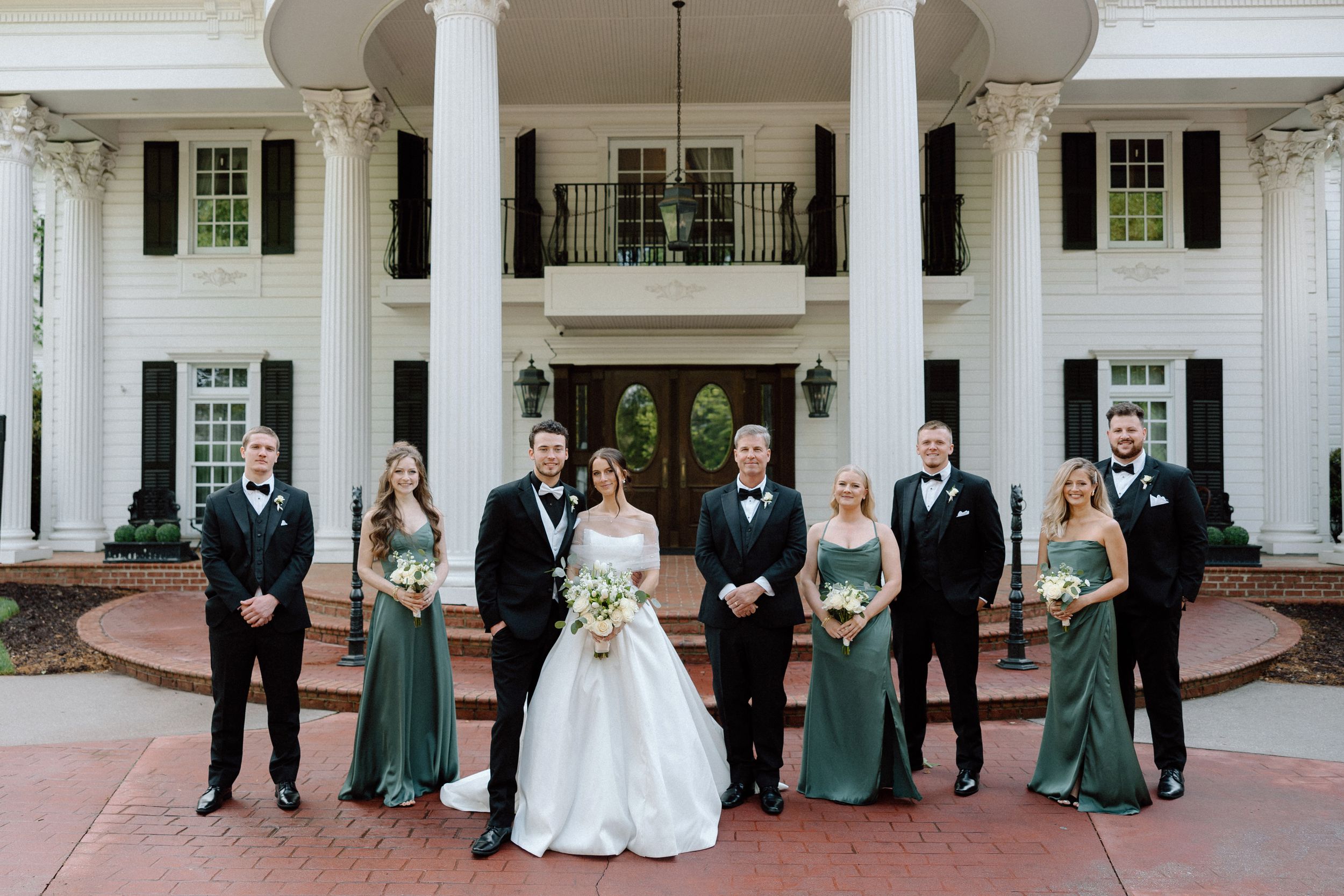 Wedding party stands in front of elegant white Southern mansion with tall columns and balcony.
