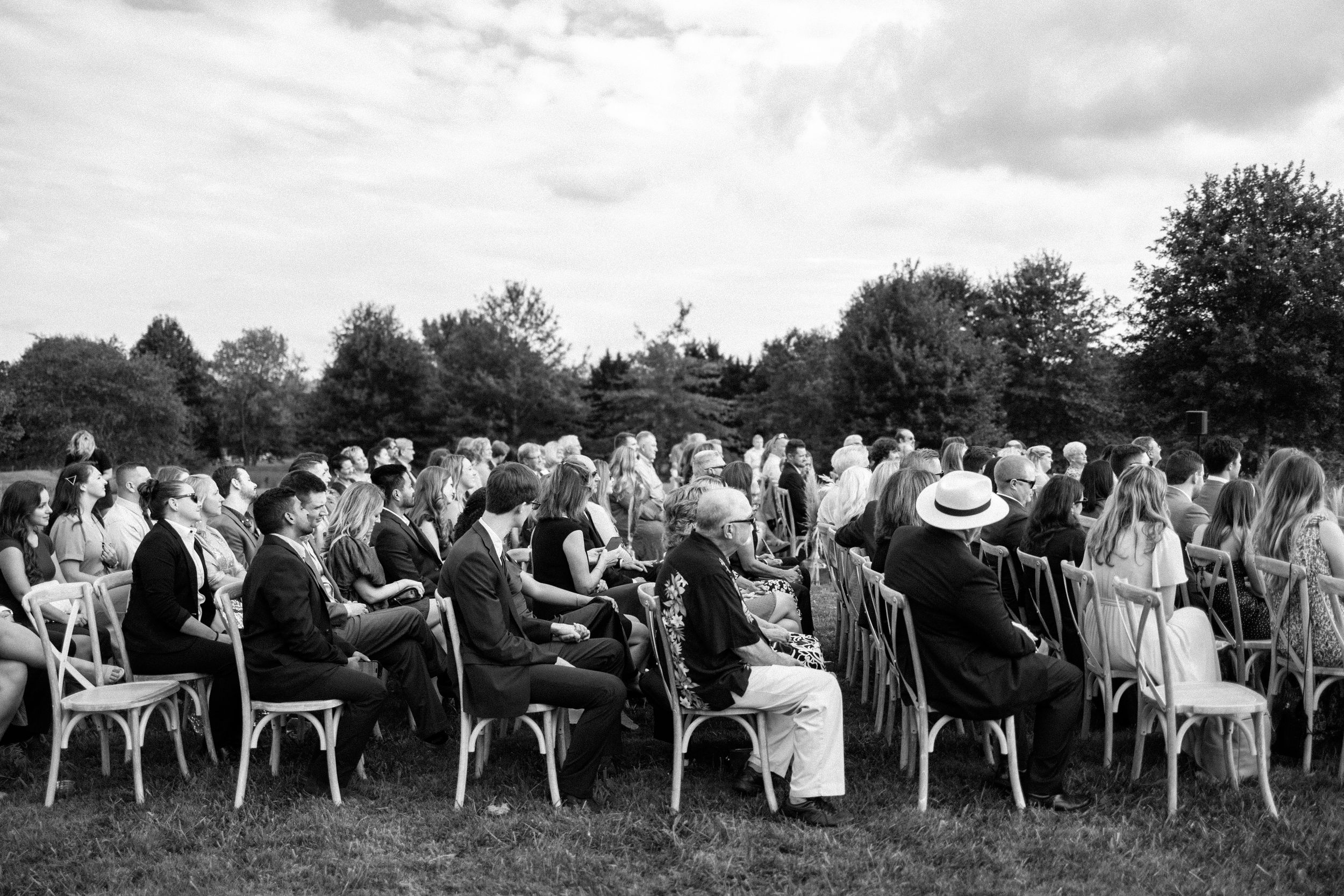 People wearing white traditional clothing stand in a line formation against a cloudy sky in a black and white photograph.