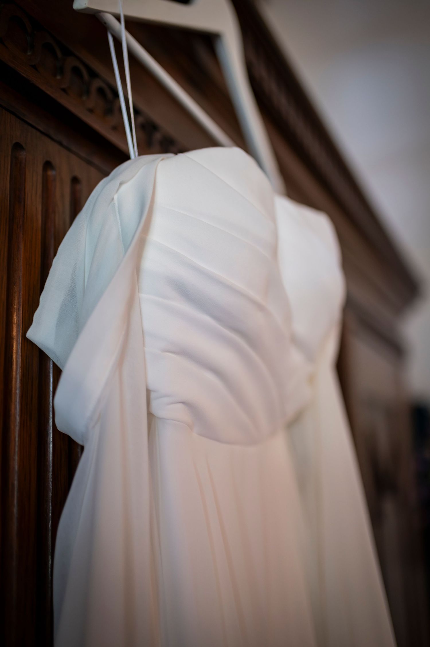 Elegant white wedding dress hangs against dark wooden panels in soft focus.