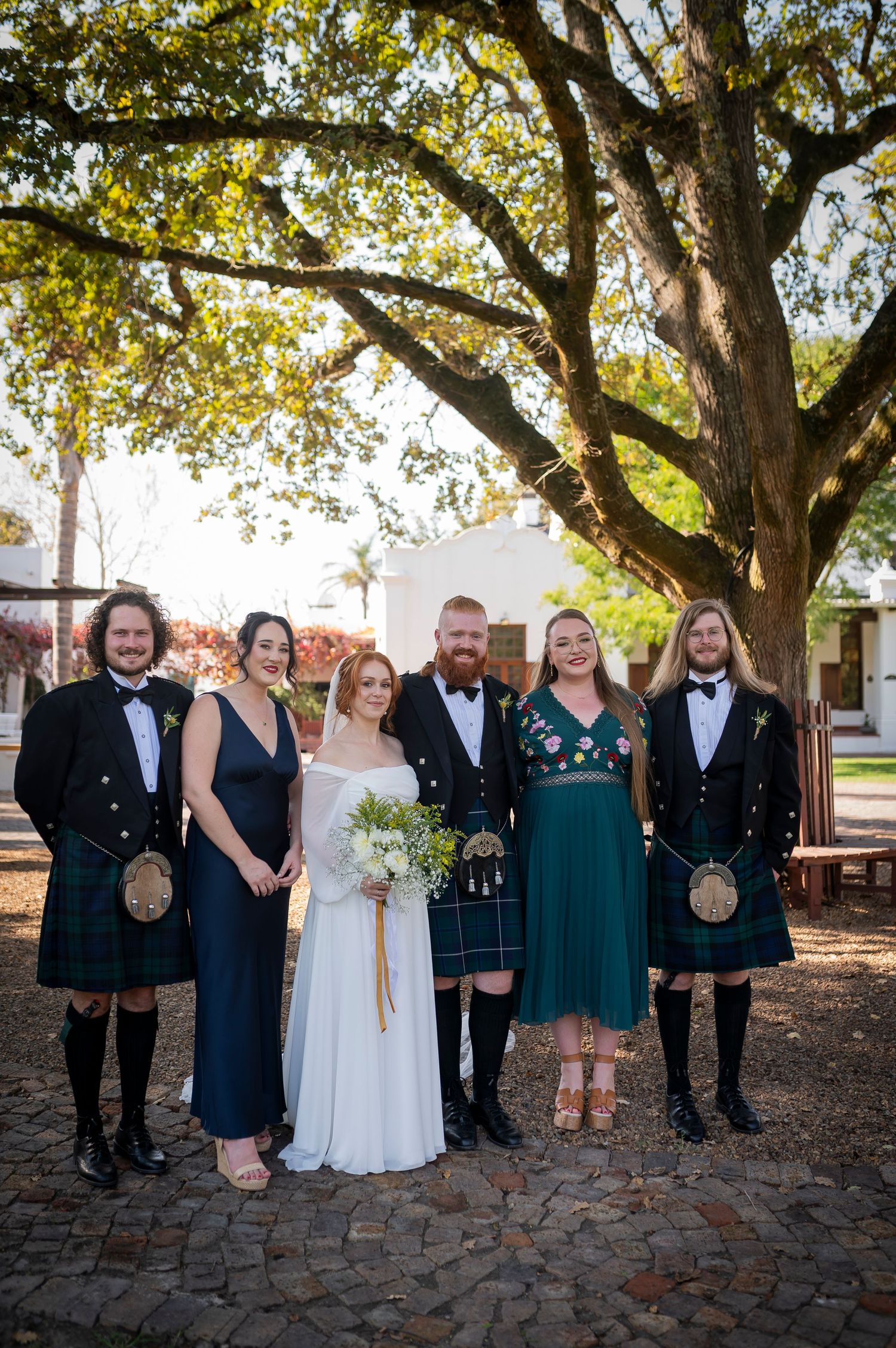 Wedding party poses outdoors under large oak tree in formal attire and traditional Scottish kilts.