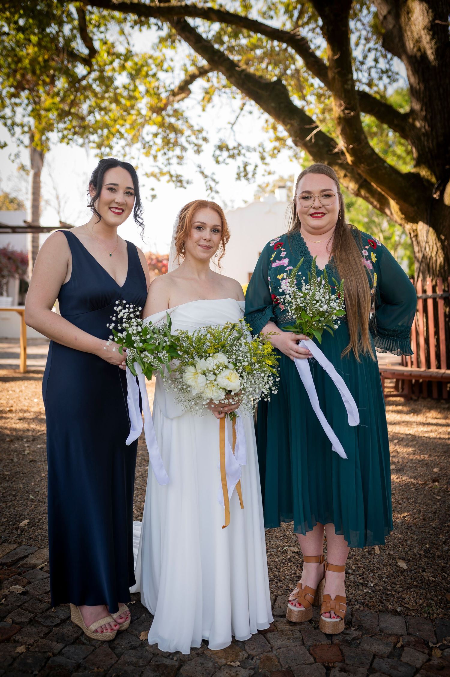 Bridal party members in navy and emerald dresses gather for photos under a sunlit tree.