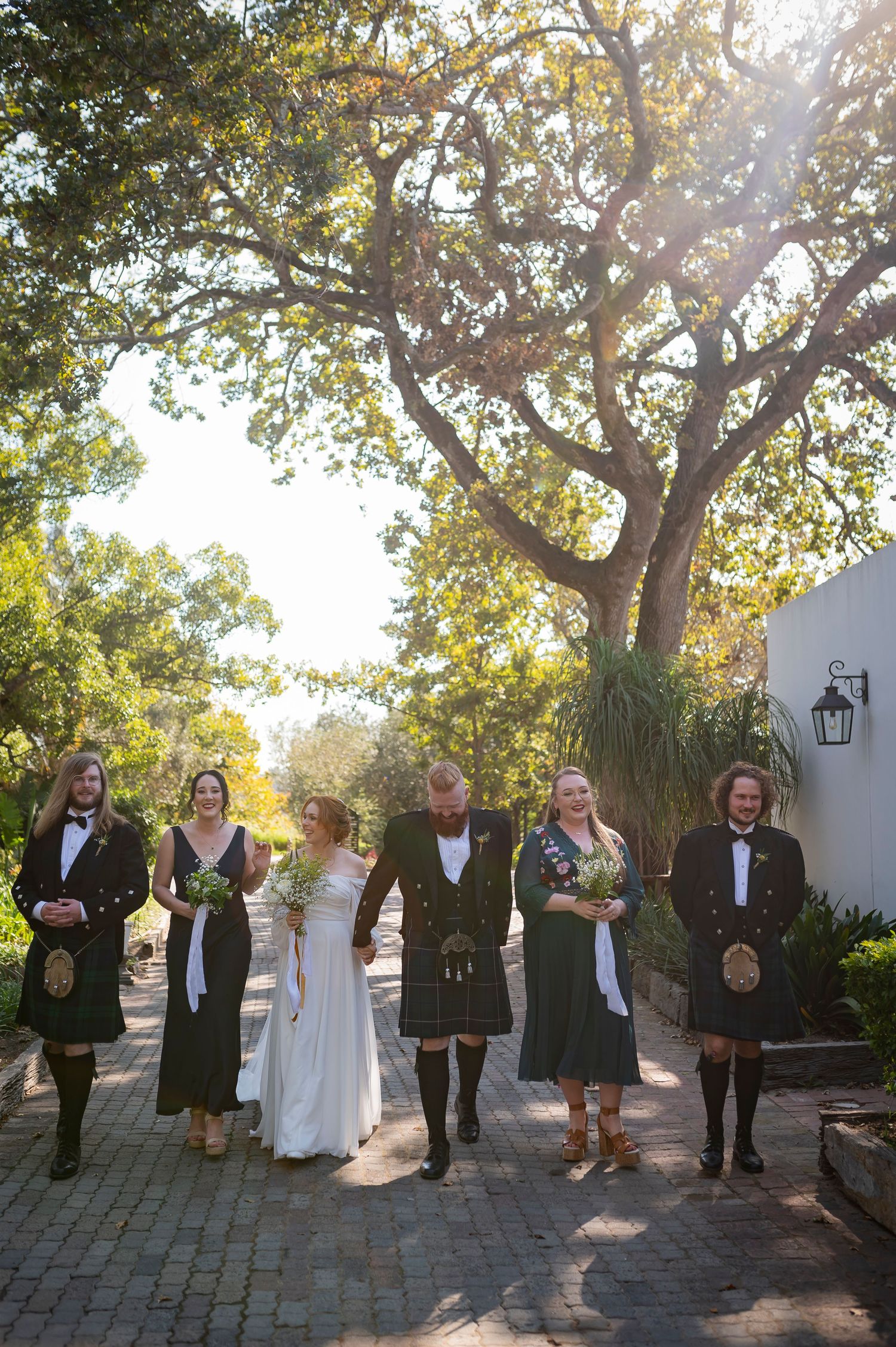 Wedding party lines up for formal photos on stone path under sunlit trees.