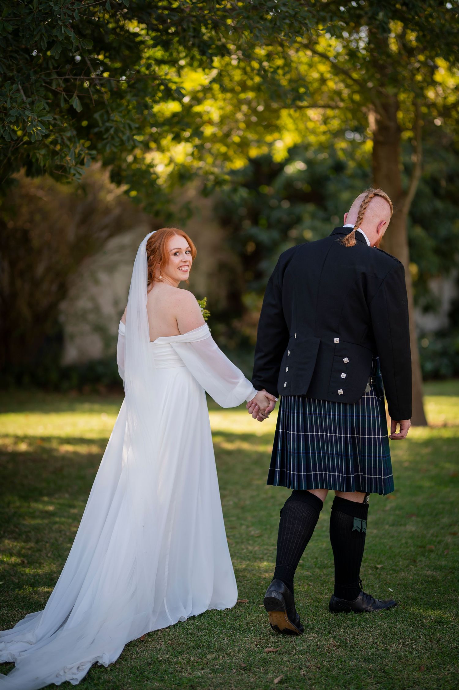 Couple poses together on grass with flowing white dress and traditional Scottish kilt.