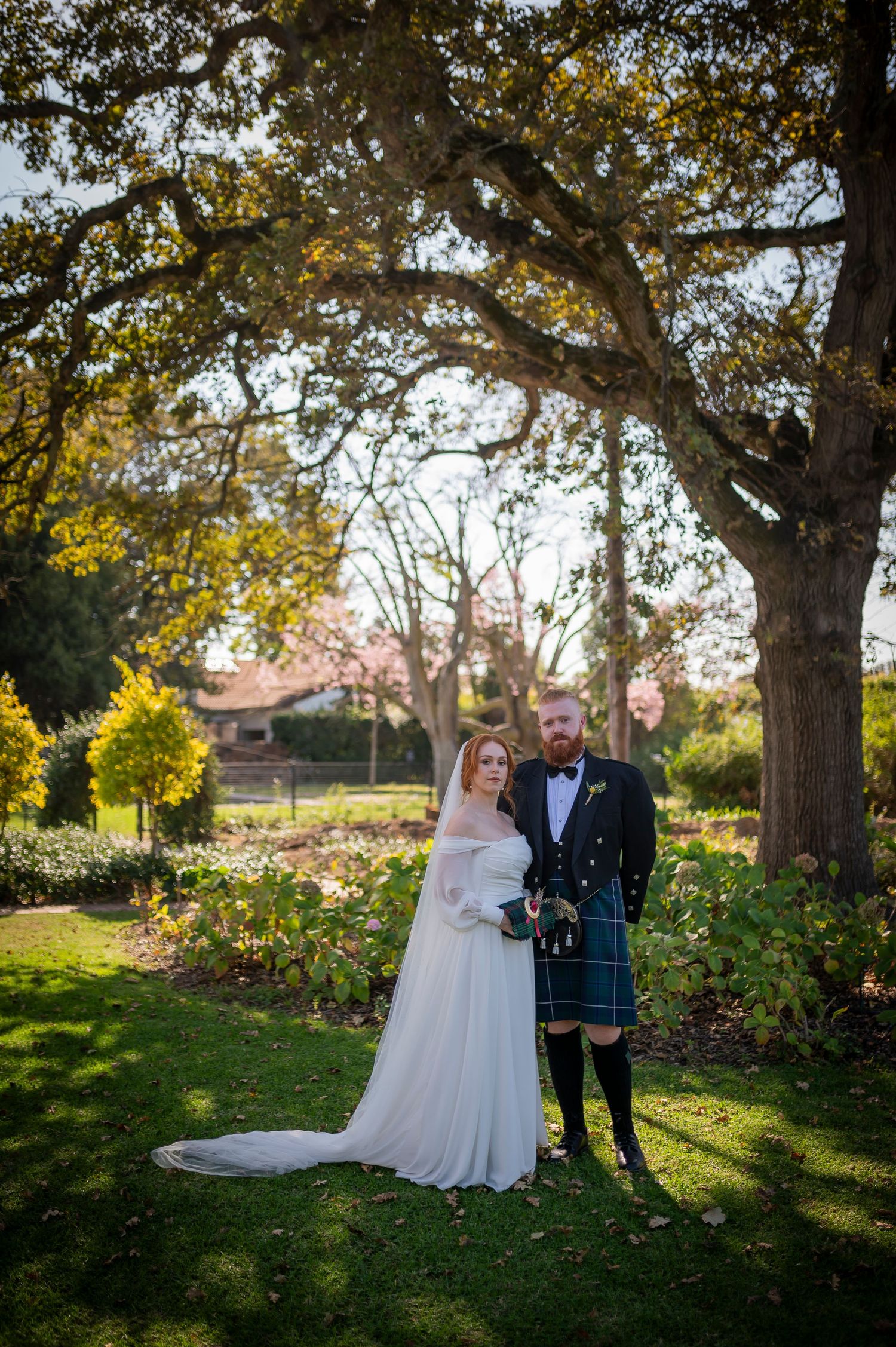 Couple poses for outdoor wedding portraits under mature oak trees in garden setting.