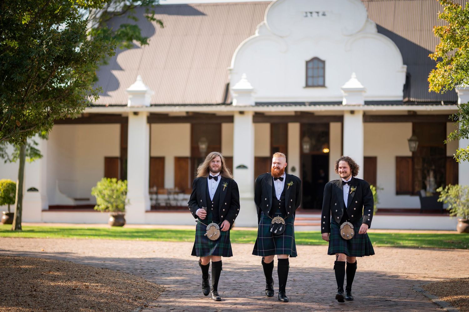 Men in formal kilts walk along white columned building exterior.