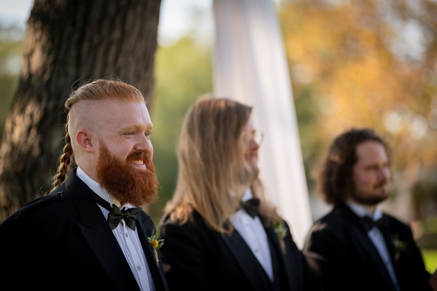 Group of men in formal attire stand together during outdoor wedding.