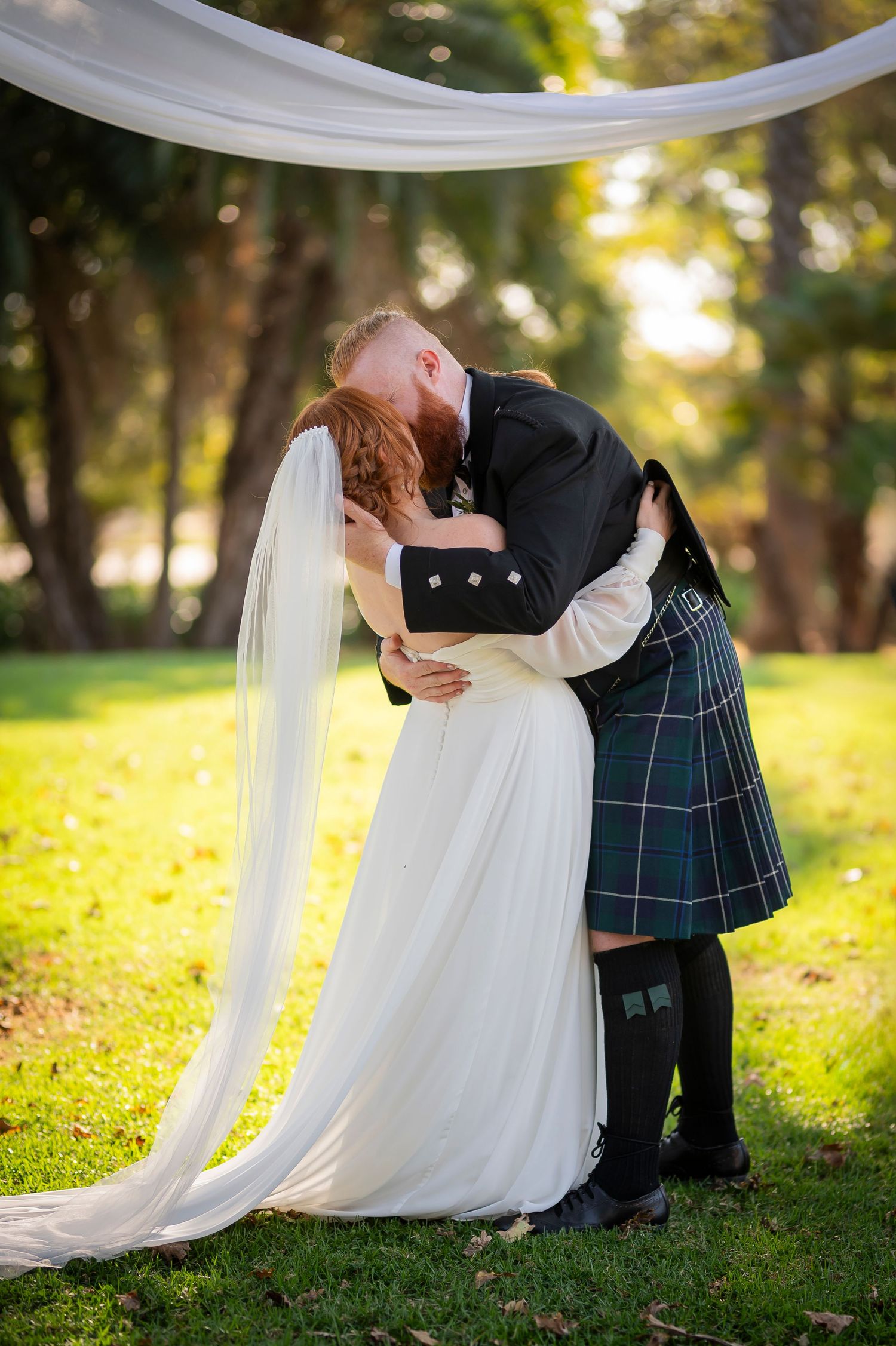 A romantic moment between newlyweds in a garden setting with the groom wearing traditional Scottish kilt.
