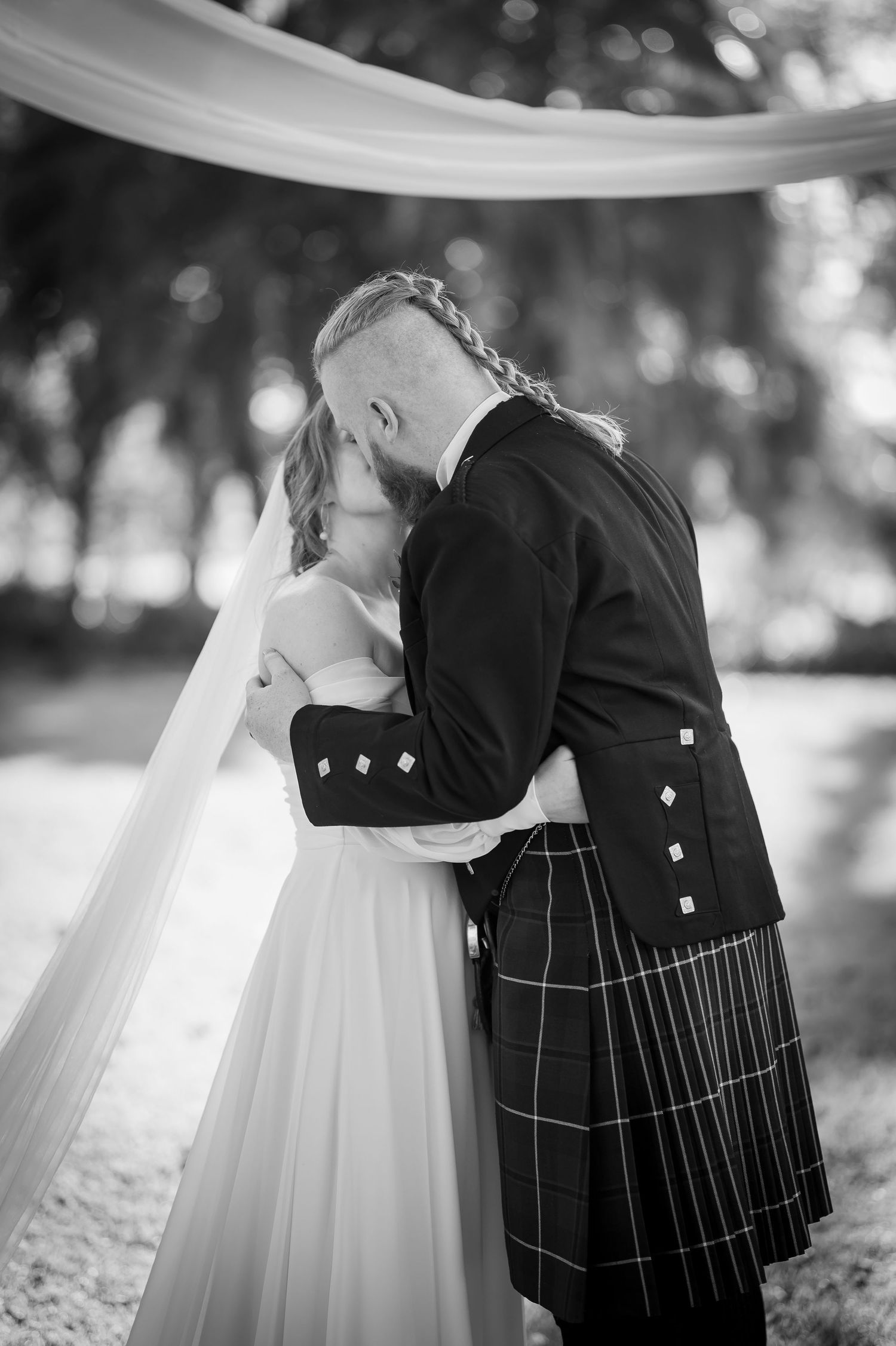 Black and white photo captures intimate moment between bride and groom in Scottish wedding attire.