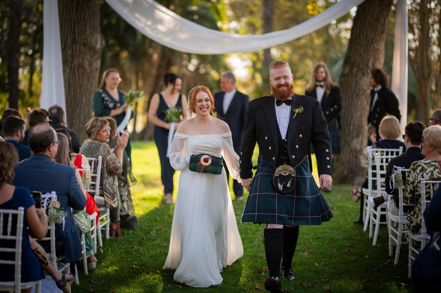 Couple walks down outdoor aisle after ceremony with guests seated in white chairs under tree canopy.