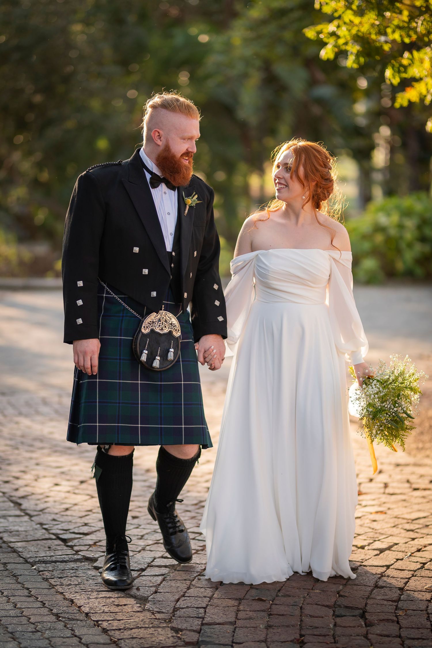 Bride in off-shoulder gown and groom in traditional kilt walk together on brick pathway at sunset.