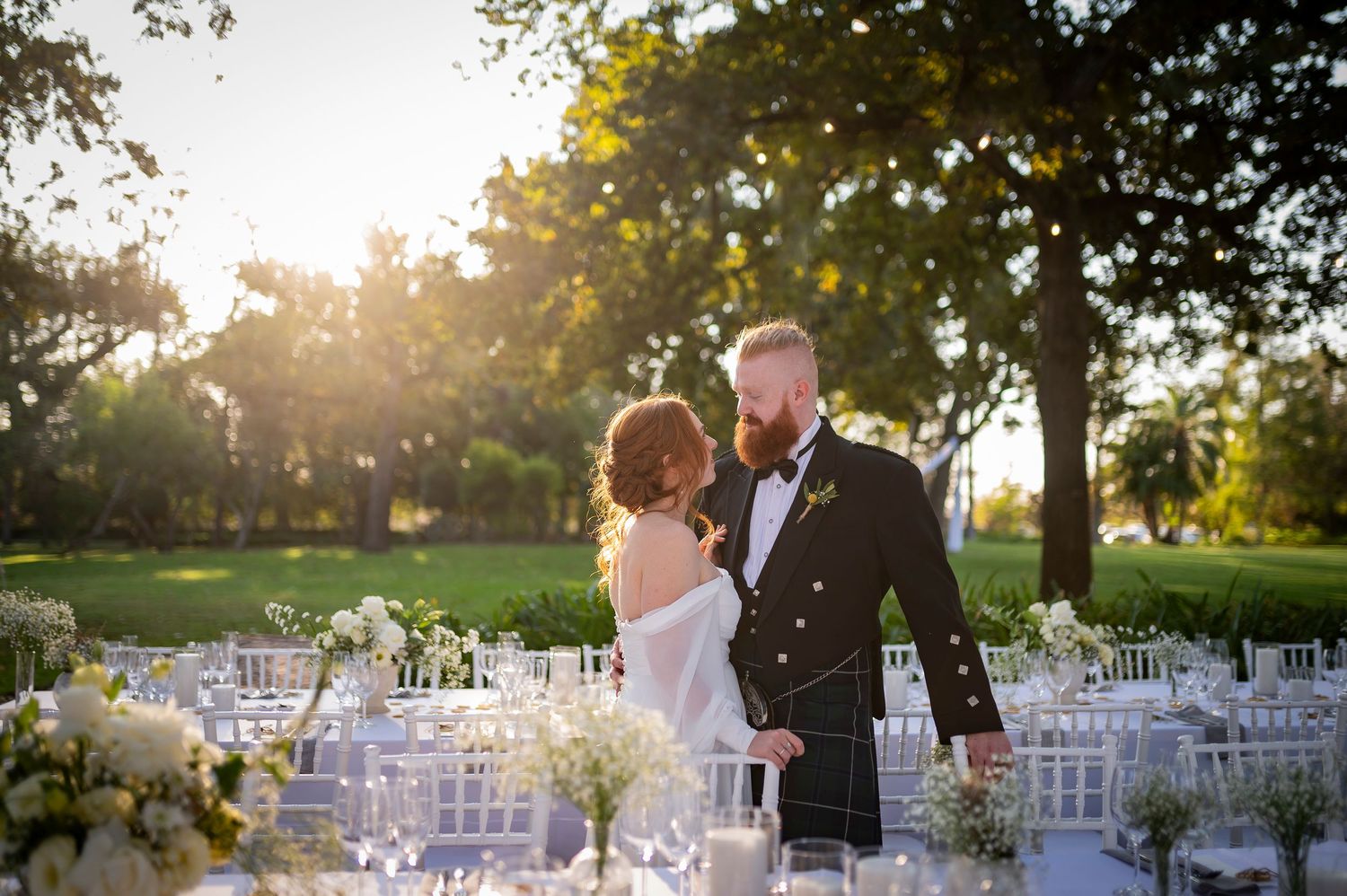 Couple admires outdoor reception setup with white chairs and elegant table settings at sunset.