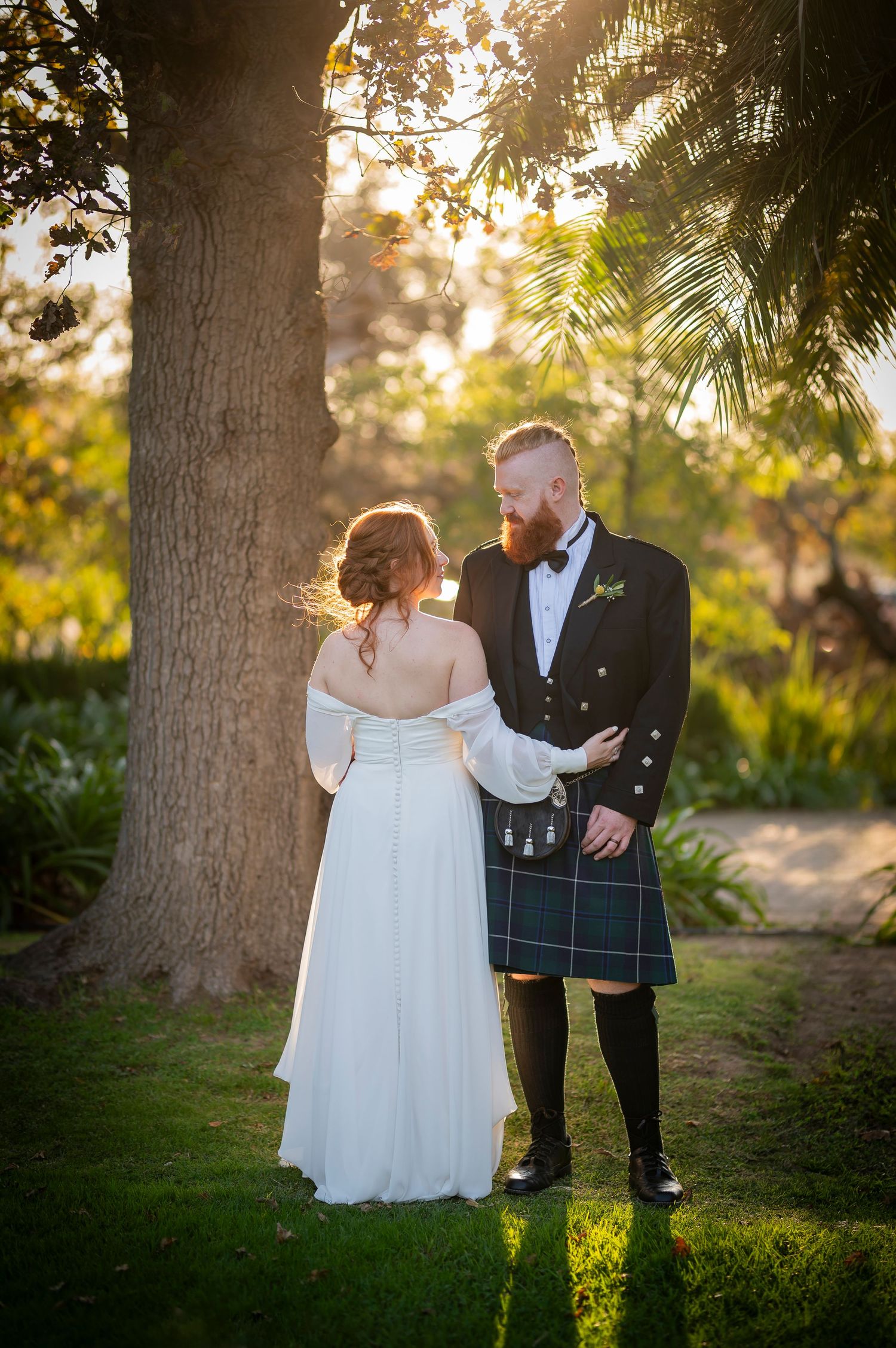 Couple shares intimate moment in Scottish wedding attire beneath a tree at sunset.