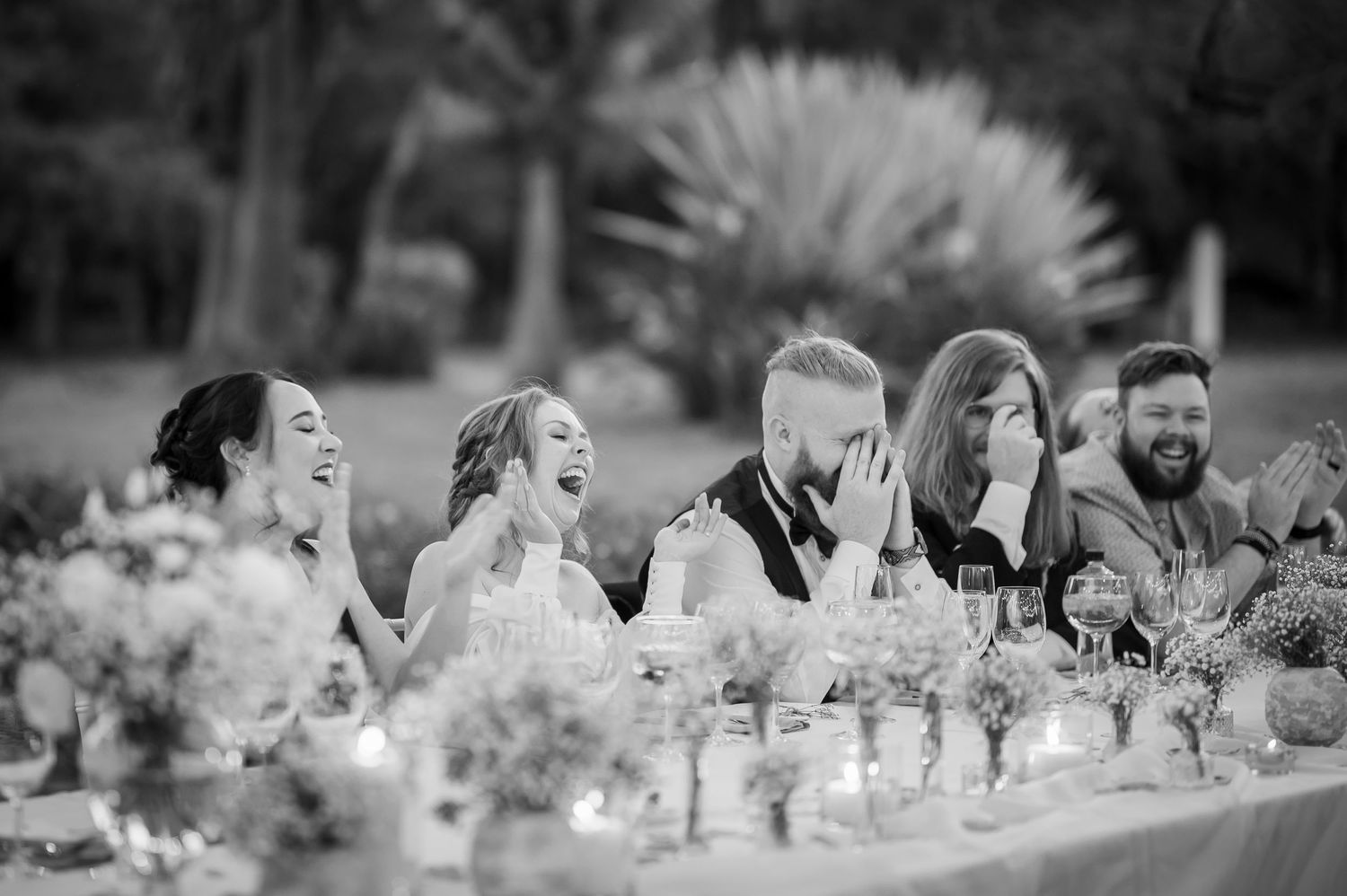 Wedding party laughing together at elegant outdoor reception with floral table decorations.