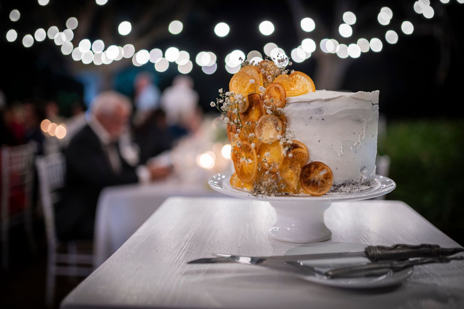 Elegant white wedding cake with golden caramel decorations under string lights.