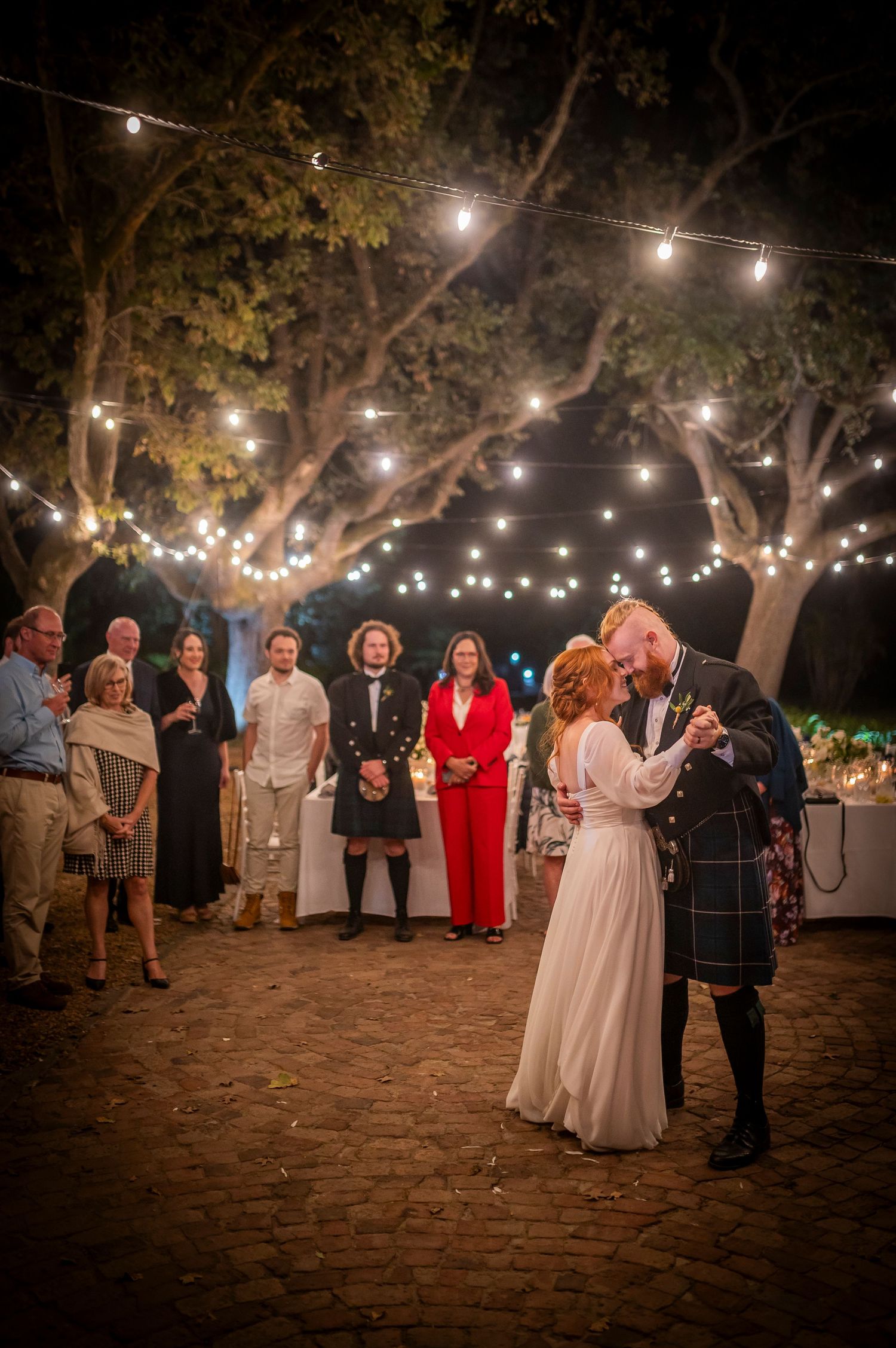 Evening wedding reception under trees with string lights as couple dances.