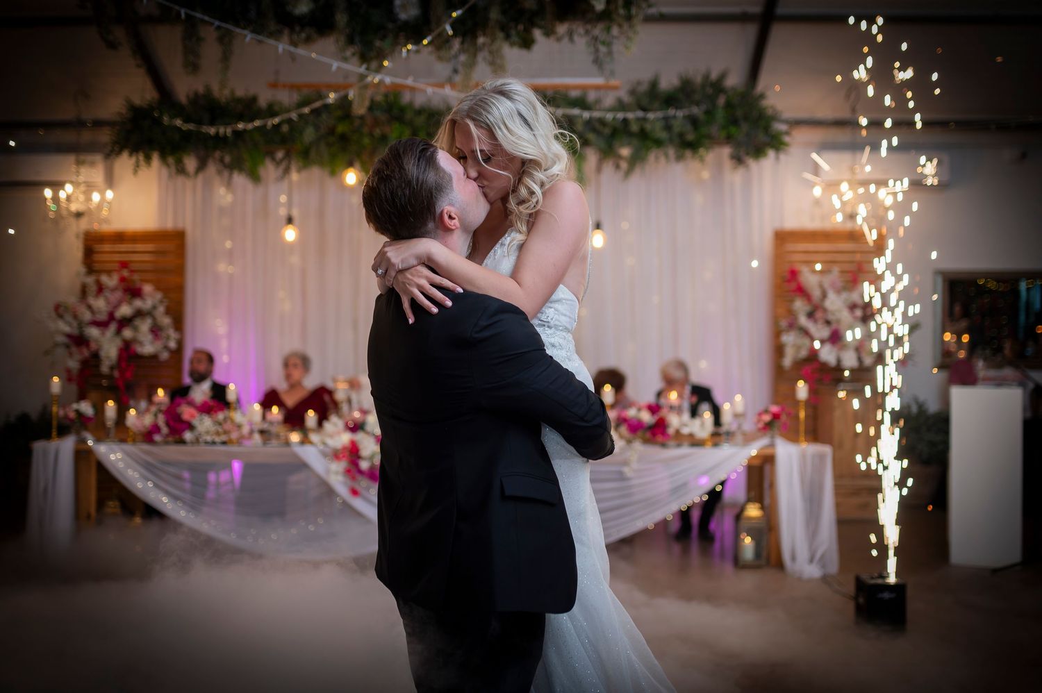 A couple shares their first dance at their wedding reception with sparklers and decorations in the background.
