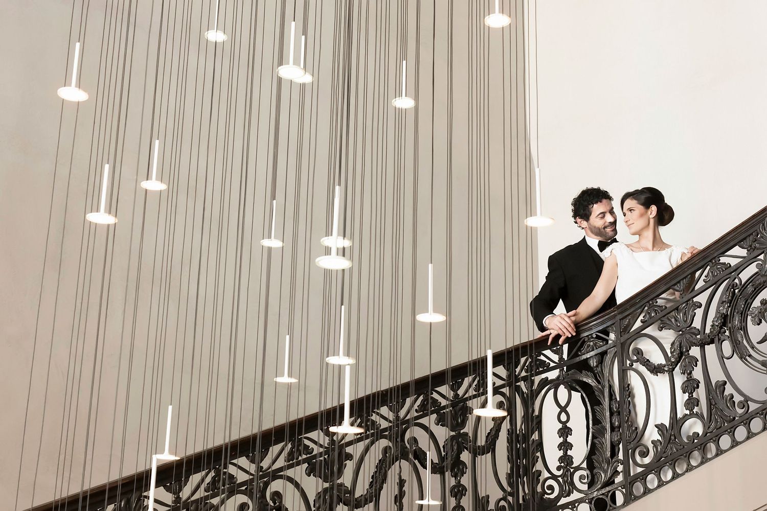 A couple stands on an ornate staircase with hanging lights and an elegant wrought iron railing.