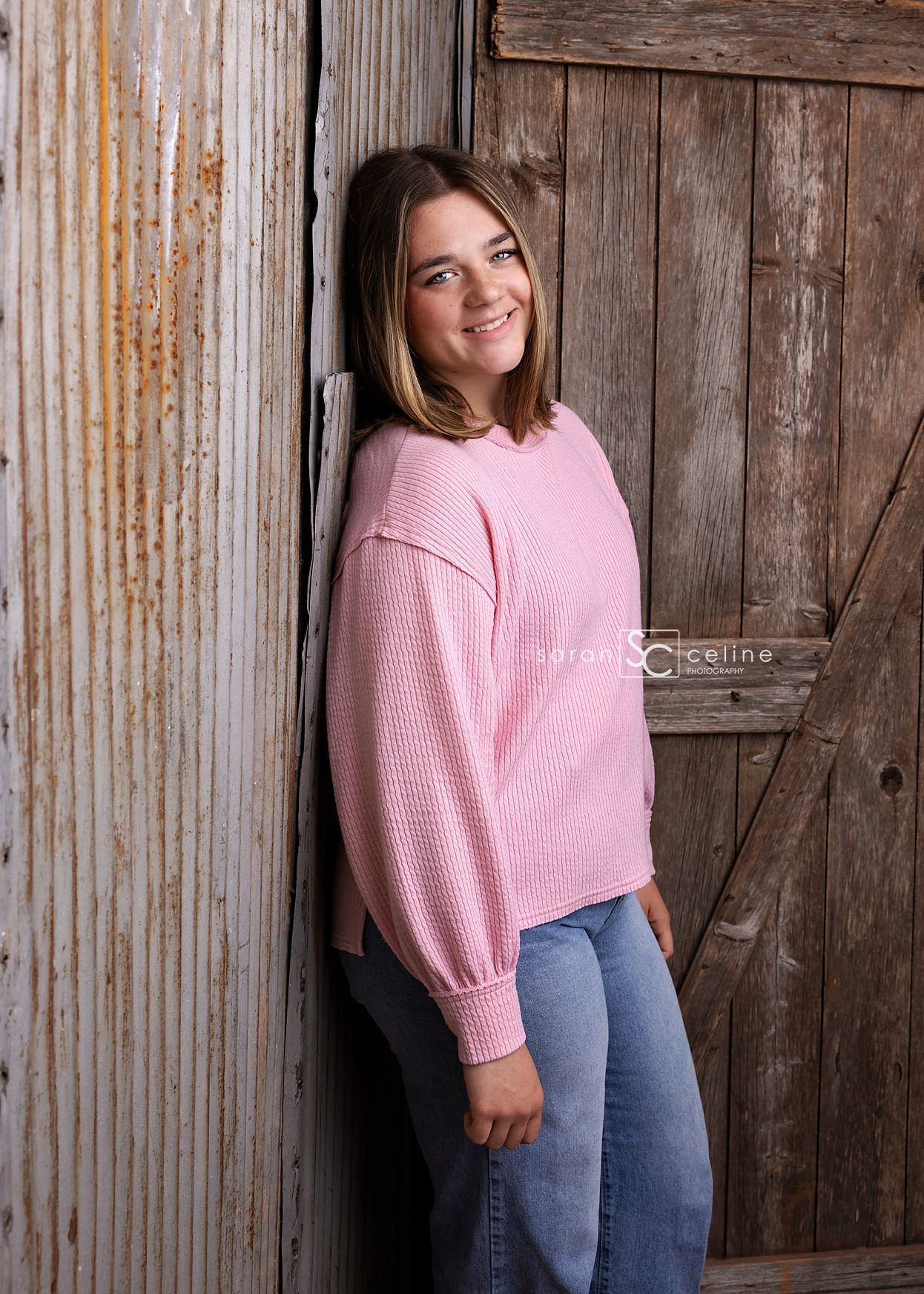 A person in a pink knit sweater and jeans poses casually against a rustic wooden barn door background.