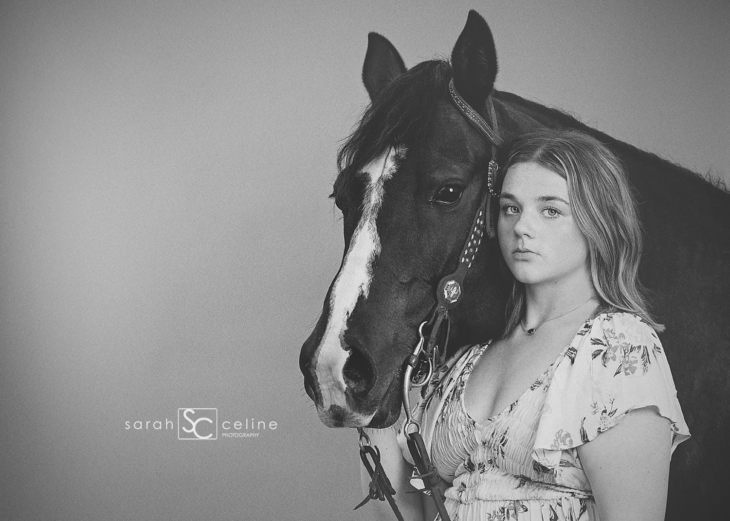 A black and white portrait of a person standing beside a dark horse with a white facial marking wearing decorative tack.