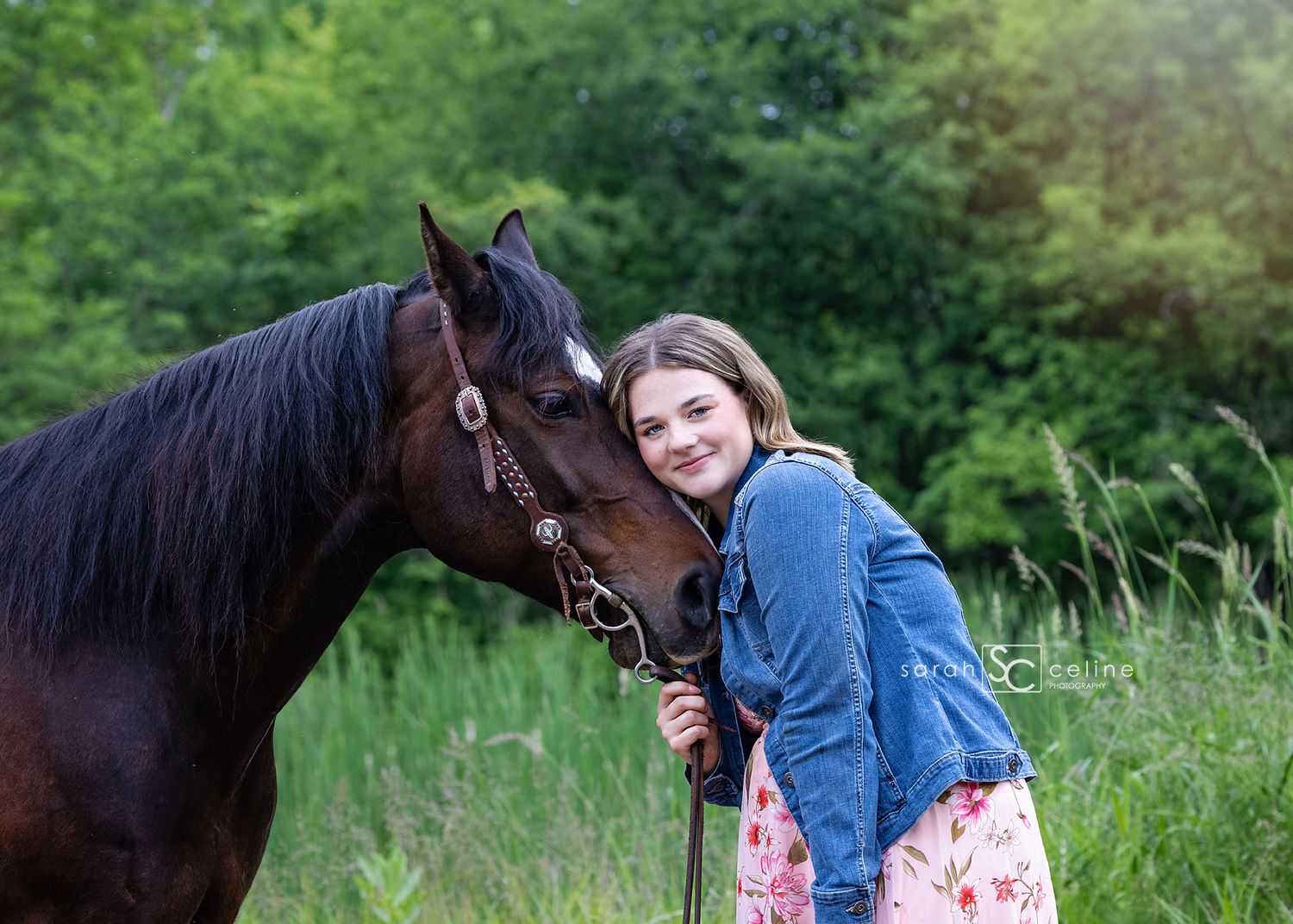 A dark brown horse and someone in a blue jacket share an affectionate moment in a lush green field.